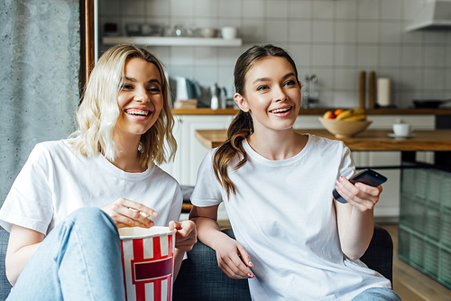 happy woman holding remote controller and watching movie with sister  near tasty popcorn