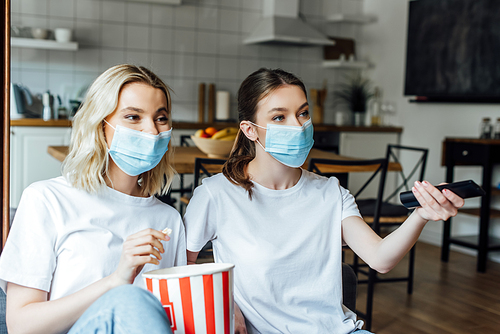 Sisters in medical masks holding popcorn while watching tv at home