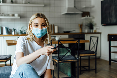 Blonde woman in medical mask holding remote controller at home