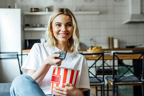 Selective focus of smiling girl holding bucket with popcorn and remote controller on couch