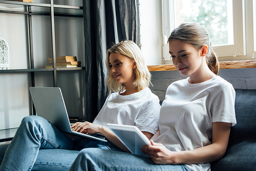 Selective focus of smiling sisters using laptop and digital tablet on sofa