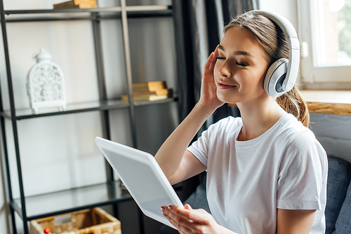 Attractive smiling woman listening music in headphones and holding digital tablet at home