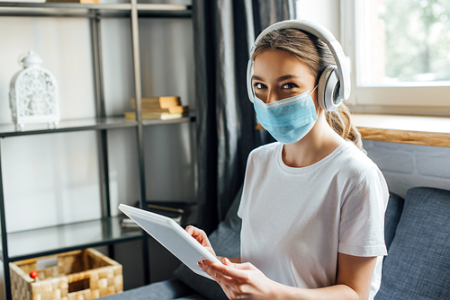 Woman in medical mask and headphones using digital tablet on couch