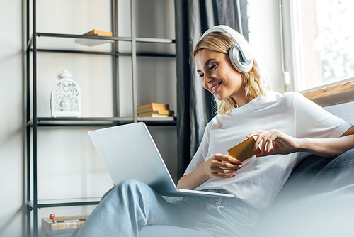 Selective focus of smiling woman in headphones using laptop and holding credit card on sofa