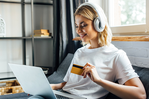 Beautiful smiling woman in headphones holding credit card and laptop on couch