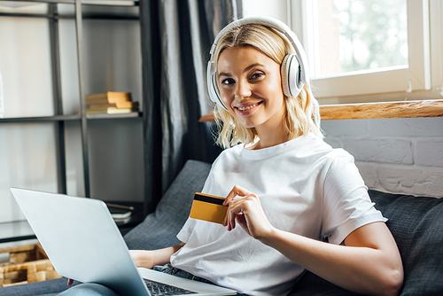 Attractive girl in headphones smiling at camera while holding credit card and laptop in living room