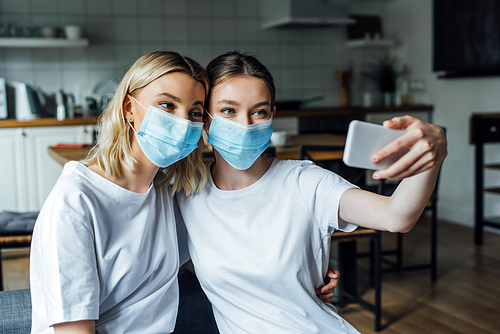 Sisters in medical masks taking selfie with smartphone at home