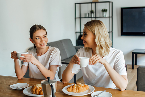 Selective focus of smiling girls drinking coffee during breakfast in kitchen