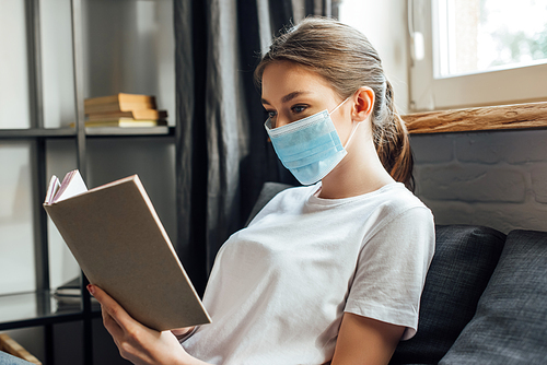 Young woman in medical mask reading book on sofa