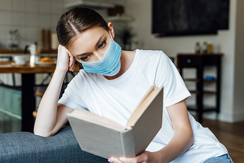 Selective focus of young woman in medical mask reading book in living room