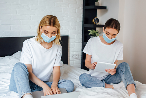 Sisters in medical masks using laptop and digital tablet on bed