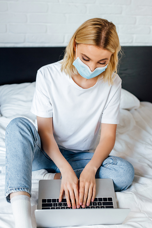 Selective focus of blonde girl in medical mask using laptop while sitting on bed