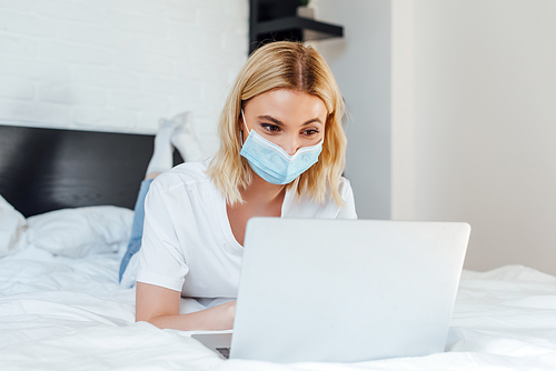 Selective focus of blonde woman in medical mask using laptop while lying on bed
