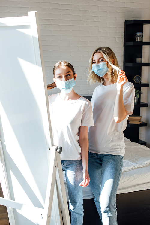 Young sisters in medical masks looking at mirror in bedroom