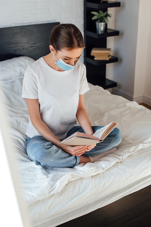 Selective focus of young woman in medical mask reading book on bed
