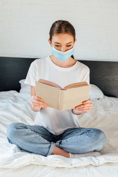Young woman in medical mask reading book while sitting with crossed legs on bed