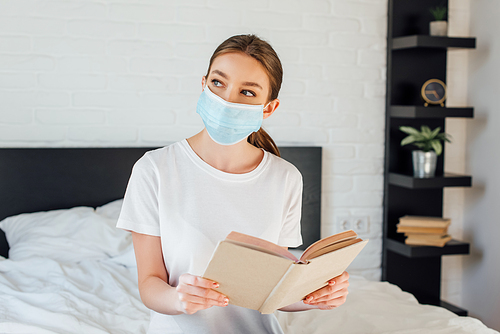 Woman in medical mask holding book while sitting on bed