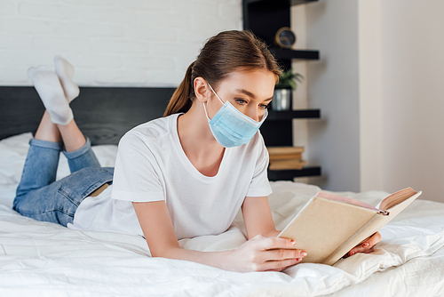 Young woman in medical mask reading book while lying on bed at home