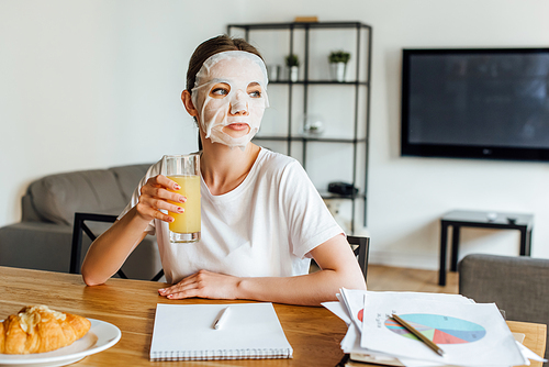 Selective focus of woman in face mask holding glass of orange juice near papers with charts on table