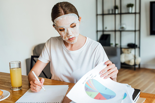 Selective focus of woman in face mask writing on notebook and holding papers with graphs near orange juice on table