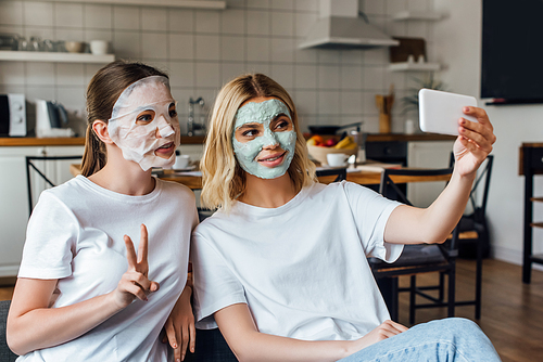 Sisters in face masks showing peace gesture and smiling while taking selfie with smartphone at home