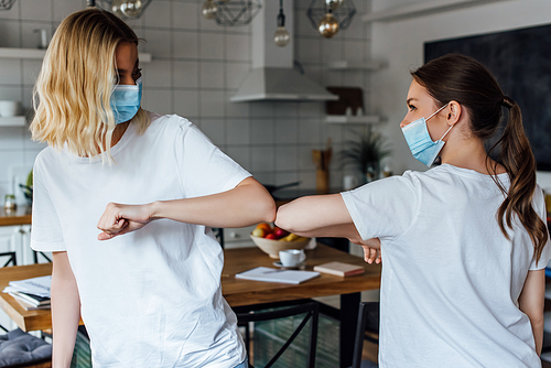 Sisters in medical masks greeting with elbow bump in living room