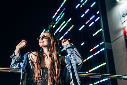 attractive woman in denim jacket and sunglasses smiling and looking away in night city