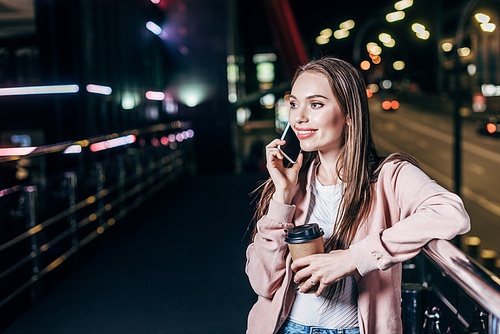 attractive woman in pink jacket talking on smartphone and holding paper cup in night city