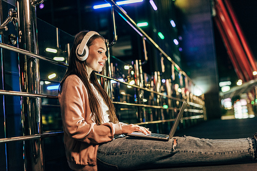 smiling woman in pink jacket with headphones using laptop in night city