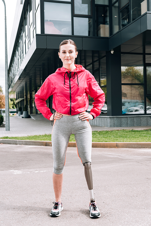 full length view of smiling disabled sportswoman standing with hands on hips on street