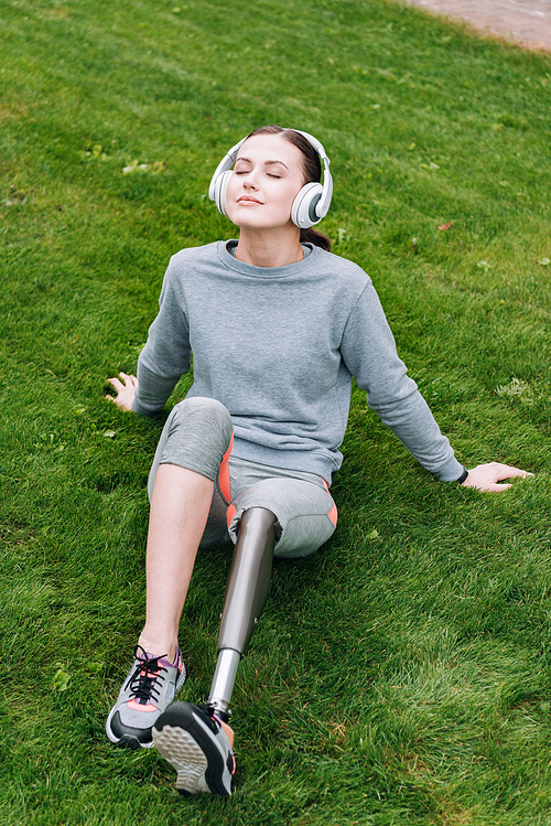 disabled sportswoman sitting on grass and listening music in headphones