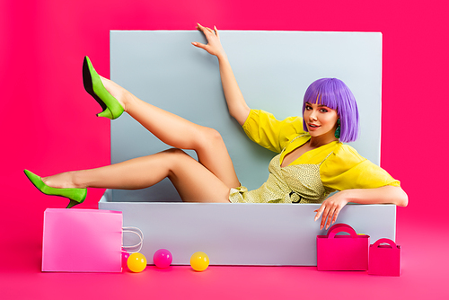beautiful emotional girl in purple wig as doll lying in blue box with shopping bags and balls, on pink
