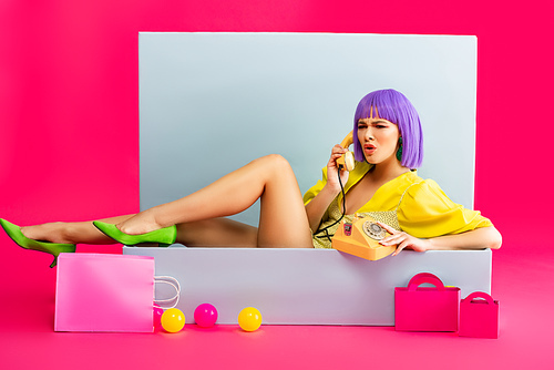 aggressive girl in purple wig as doll talking on vintage phone while sitting in blue box with balls and shopping bags, on pink