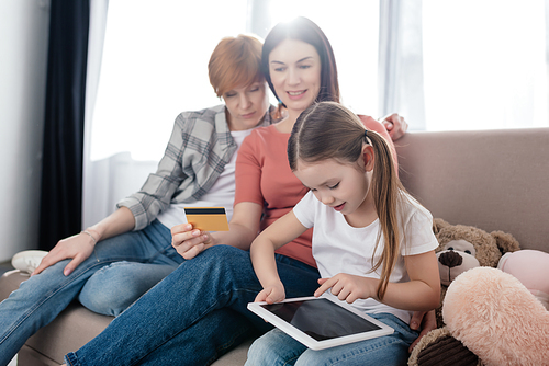 Selective focus of kid using digital tablet near mothers with credit card on couch in living room