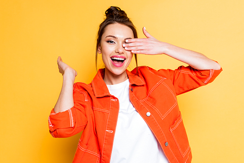 cheerful young woman covering eye with hand while smiling at camera on yellow background