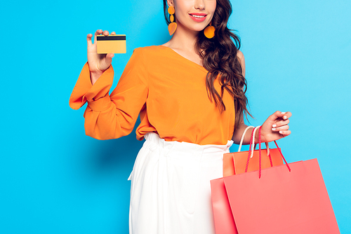 cropped view of a fashionable girl holding shopping bags and showing credit card on blue background