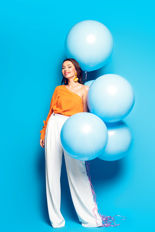 full length view of cheerful stylish woman with big festive balloons smiling at camera on blue background