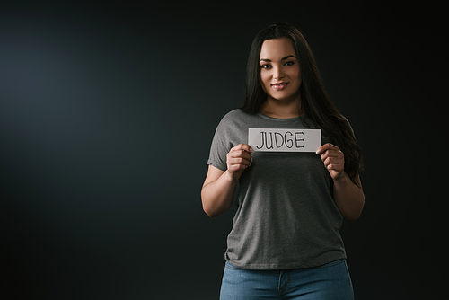 Front view of smiling plus size girl holding card with word judge on black background