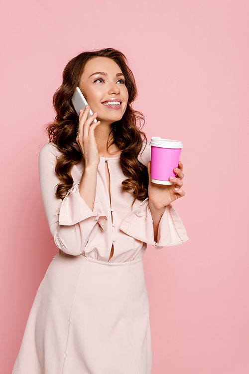 happy girl talking on smartphone and holding paper cup isolated on pink