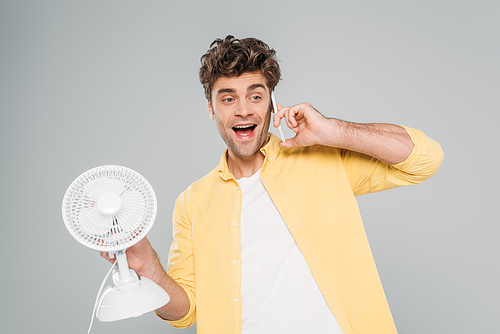 Front view of excited man holding desk fan and talking on smartphone isolated on grey