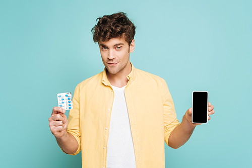 Front view of man smiling,  and showing smartphone and blister pack with pills isolated on blue