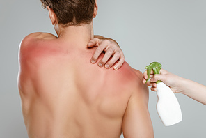 Cropped view of woman with bottle of spray near man touching skin with sunburn isolated on grey