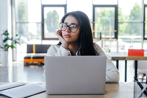 sad businesswoman in glasses looking away near laptop and notebook on table