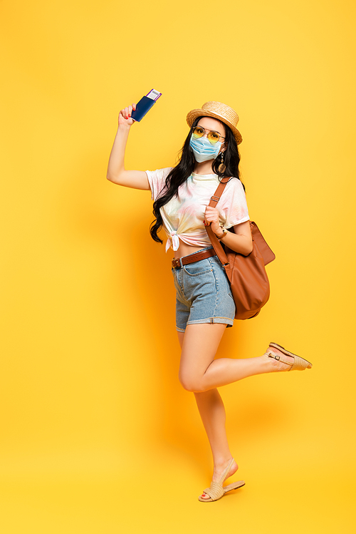 brunette girl in summer outfit and medical mask with bag and boarding pass on yellow background