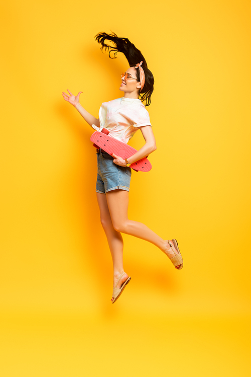 side view of happy stylish summer brunette girl jumping with pink penny board on yellow background