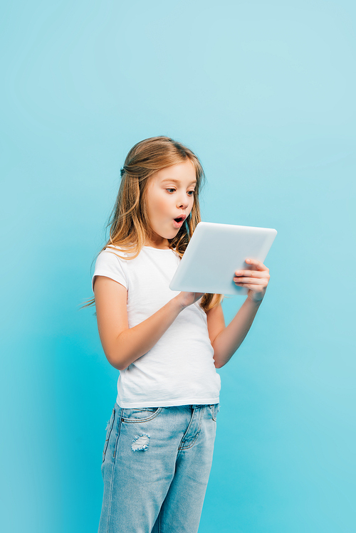 excited child in white t-shirt and jeans using digital tablet isolated on blue