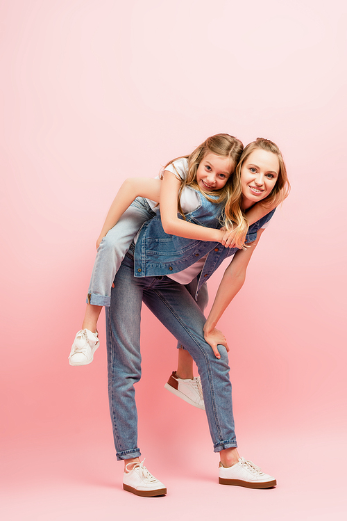 full length view of young woman in denim clothes piggybacking kid on pink