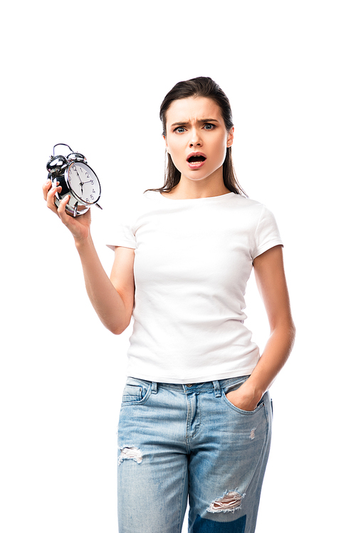 shocked woman in white t-shirt and jeans standing with hand in pocket and holding retro alarm clock isolated on white