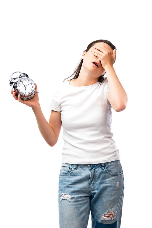 tired woman in white t-shirt and jeans holding retro alarm clock while covering face isolated on white