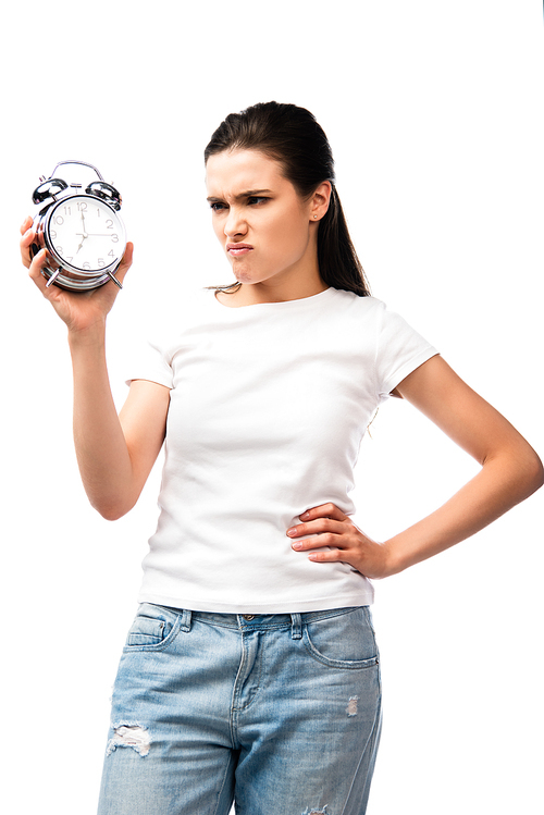 displeased woman in white t-shirt holding retro alarm clock while standing with hand on hip isolated on white